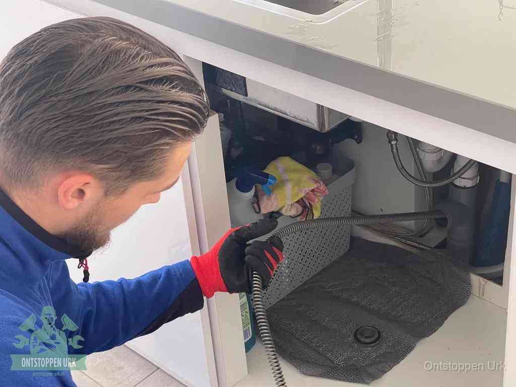 Een monteur voert een flexibele ontstoppingsveer in onder het keukenkastje, langs de sifon, om de gootsteen te ontstoppen in Urk