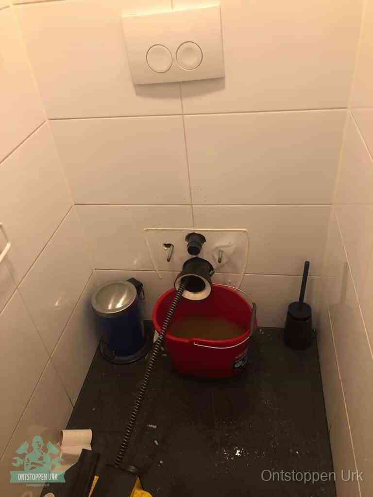 Een ontstopte wand-wc met verwijderde toiletpot waar een ontstoppingsveer in de wanduitgang verdwijnt, het achterhalen waar de verstopping zit in Urk