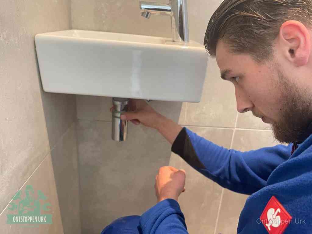 Een monteur draait met de hand de zwanenhalssifon onder een fonteinwasbak in de toiletruimte los om de bron van rioollucht in Urk te vinden