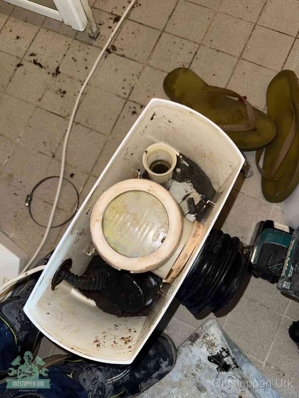 Een gedemonteerde toiletpot op de vloer met ontstoppingsveer en gereedschap ernaast, het verwijderen van een vastzittend voorwerp uit de wc in Urk